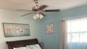 Quarto com cama, mesa de cabeceira, cadeira, ventoinha no teto, pintura e janela com cortinas.
