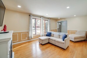 A living room with a beige sectional sofa, a small ottoman, a flat-screen TV, and a kitchen area with a red appliance. - 1 Block to Red Line Station: Walkable Chicago Home (Chicago)