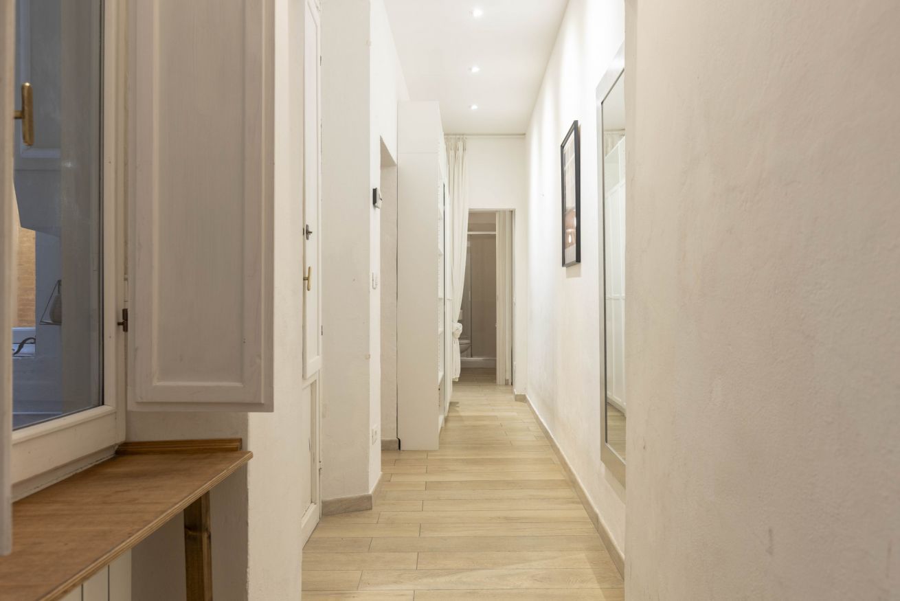 Un couloir étroit aux murs blancs et au plancher en bois, qui mène à une pièce dotée d’une fenêtre et d’une porte.