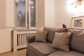 A living room with a grey sofa, two patterned cushions, a white radiator, and a window with a view of greenery. - Ghibellina Comfort Apartment (Firenze)