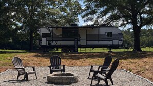 A campsite with a camper RV, a fire pit, and Adirondack chairs.