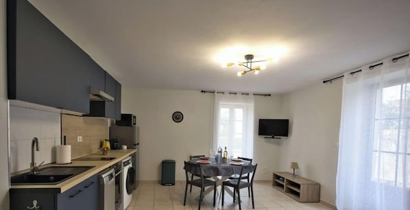 A compact kitchen with a dining area, a TV mounted on the wall, and a window with curtains. - Hostcare - La Petite Escale: Parking (La Rochelle)