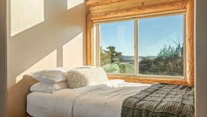Un dormitorio con un ventanal grande, una cama con ropa de cama blanca y vistas a un paisaje boscoso.