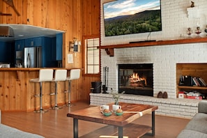 A cozy living room with a fireplace, wooden walls, a kitchen area, and a bar with stools. - Solstice Shores Chalet, Hot Tub and Lake Views (Fawnskin)