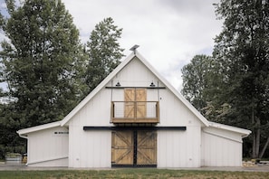 A white barn-style building with a gabled roof, a wooden door, and a small balcony with a railing. - Riverfront Barndominium, Incredible Outdoor Space! (Townsend)