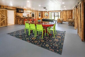 A dining area with a wooden table and green chairs, a bar area with a TV, and a large window. - Riverfront Barndominium, Incredible Outdoor Space! (Townsend)