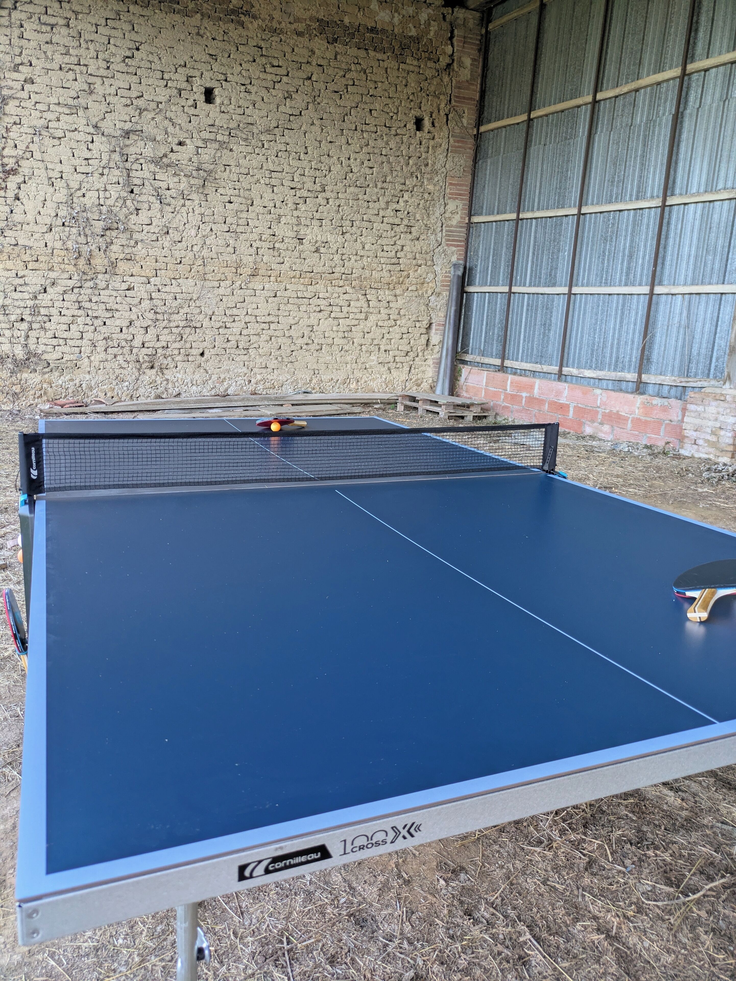 Mesa de ping pong con red en un área exterior con pared de ladrillo y cerca de metal corrugado al fondo.