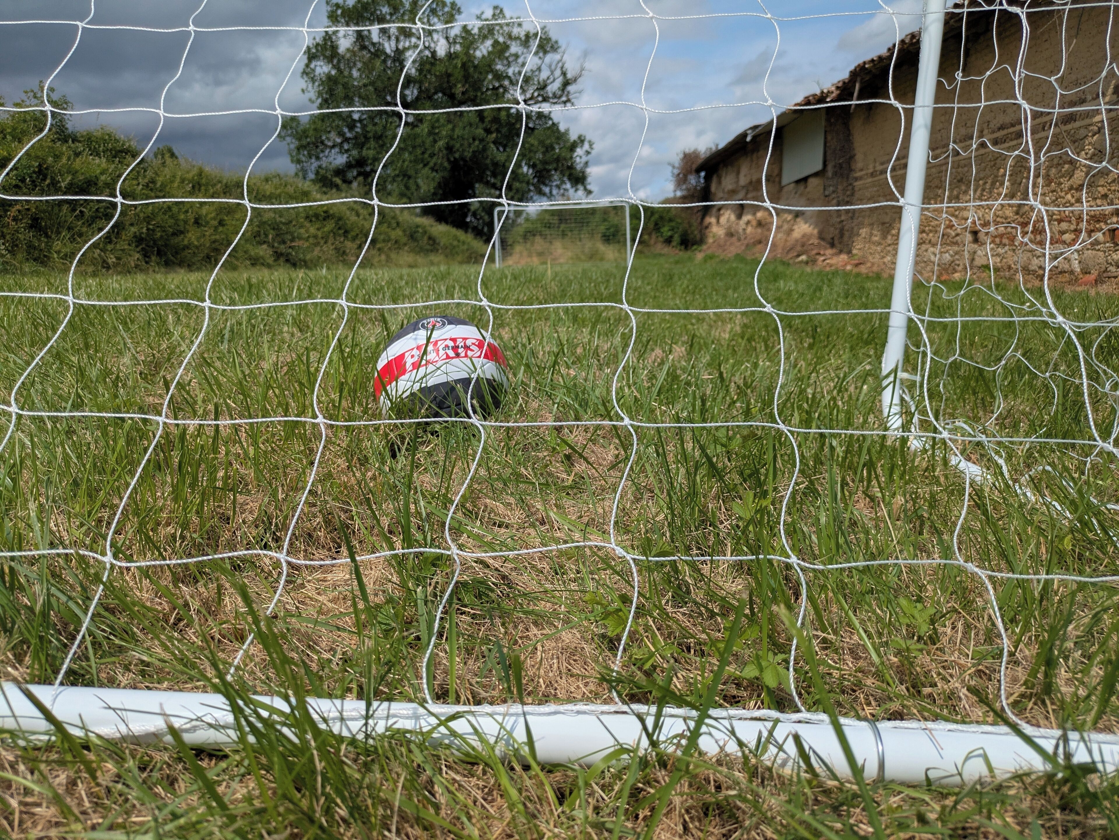 Un ballon de football près d’une clôture en fil de fer, avec de l’herbe et un bâtiment en arrière-plan.