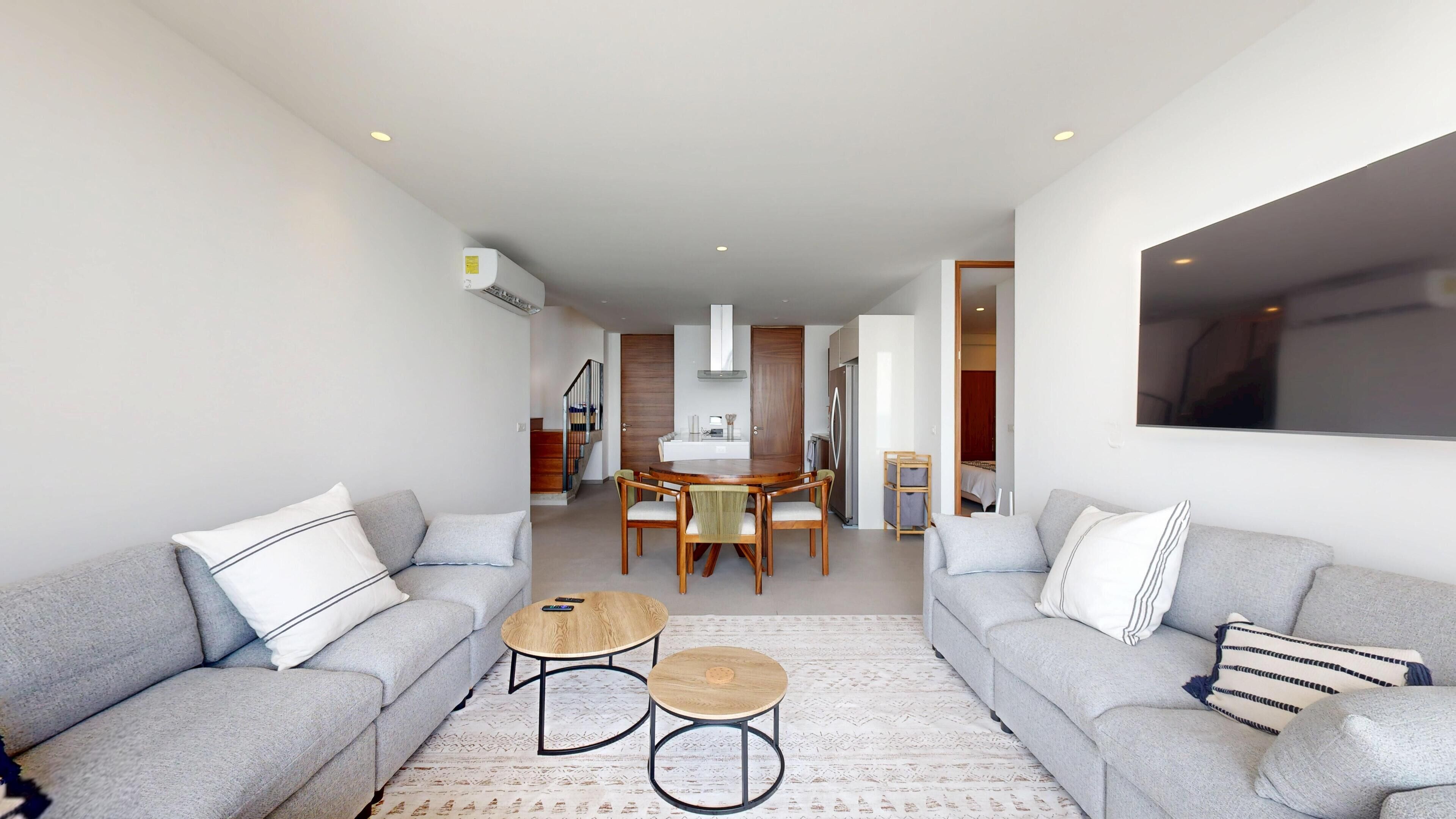 Uma sala de estar moderna com dois sofás cinzas, uma mesa de centro e uma área de jantar com mesa e cadeiras de madeira.