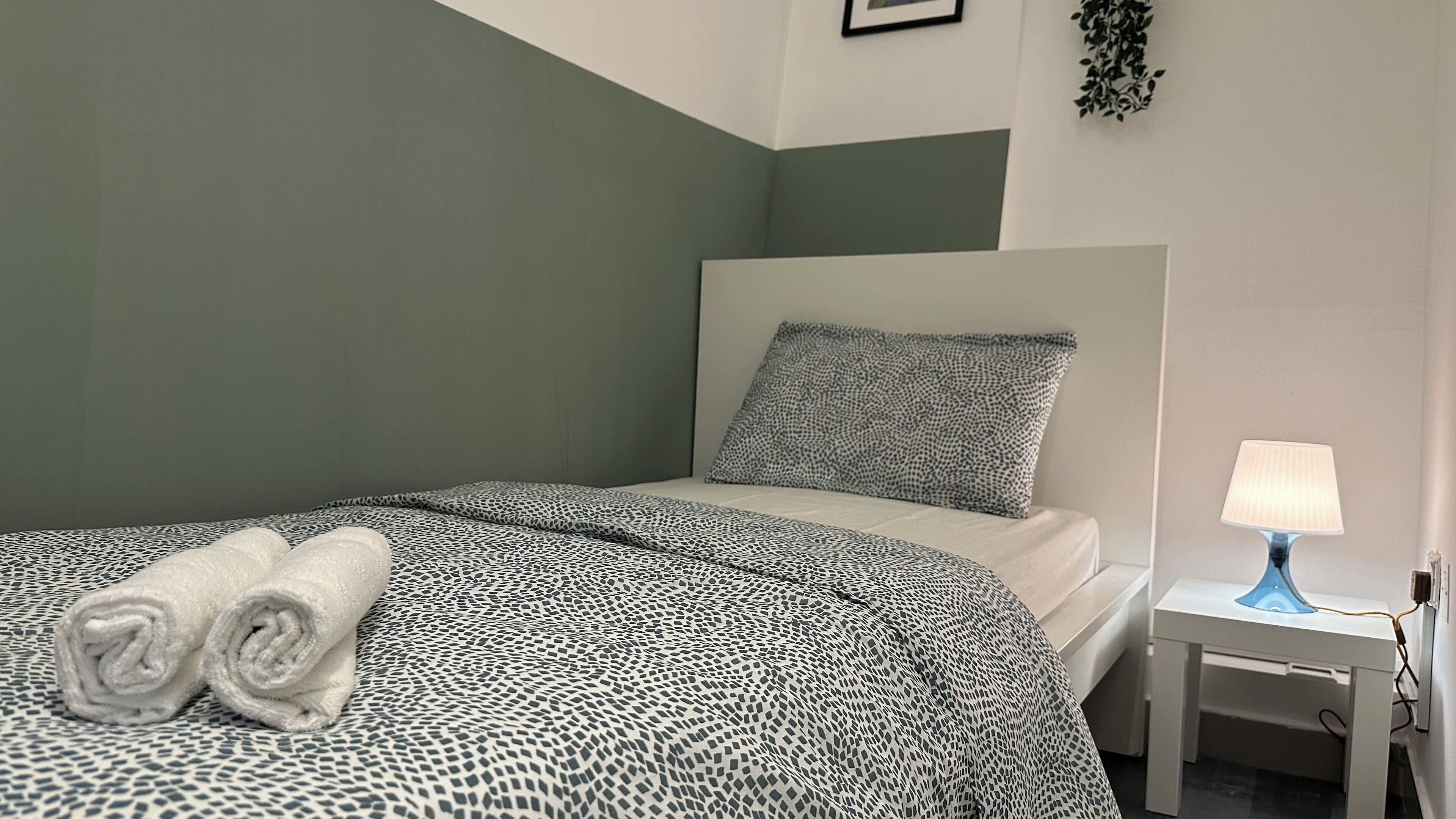 Un lit bien fait, avec une couvre-lit à motifs et des oreillers, une table de chevet avec une lampe et une plante fixée au mur.