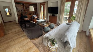 Un salón amplio con un sofá, sillones, una mesa de centro y un televisor. Hay una estantería llena de libros y objetos decorativos.