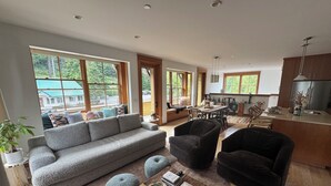 Amplia sala de estar con sofá gris, sillones negros y una mesa de centro con libros y objetos decorativos. Amplias ventanas ofrecen vistas al exterior.