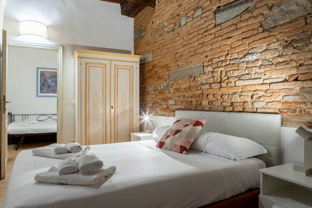A hotel room with a brick wall, a bed with white linens, a nightstand, and a wooden wardrobe.
