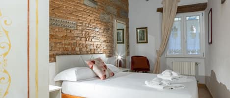 Un dormitorio con pared de ladrillo, cama con ropa blanca, una silla roja y ventana con cortinas.