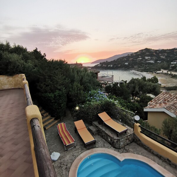 Área de piscina com espreguiçadeiras e vista para o pôr do sol sobre a paisagem costeira.