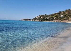 Uma praia com águas azul-claras, areal arenoso e casas numa encosta.