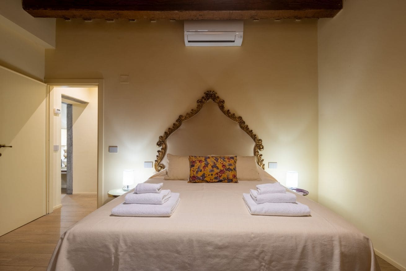 A hotel room with a large bed, two bedside tables, an air conditioning unit, and a door leading to another room.