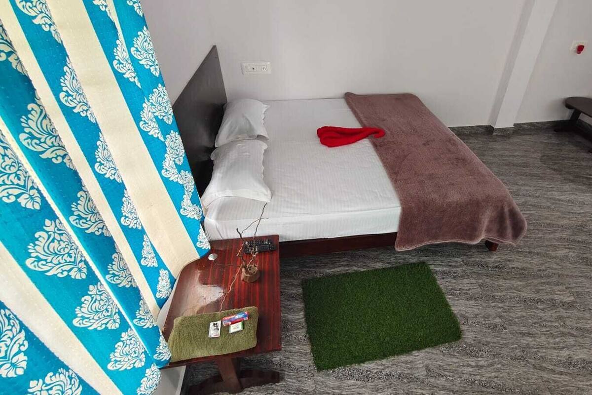 A small, neatly arranged room with a bed, a bench, a green rug, and a small table with a bottle and a tissue box.