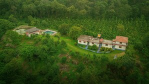 Una casa grande de dos pisos rodeada de un bosque frondoso, con piscina y un pequeño jardín.