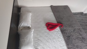 Una cama bien hecha con ropa blanca, una manta gris y una toalla roja.