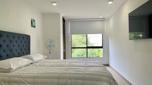 Un dormitorio moderno con una cama grande, un ventilador de pie y un ventanal con vistas a zonas verdes.