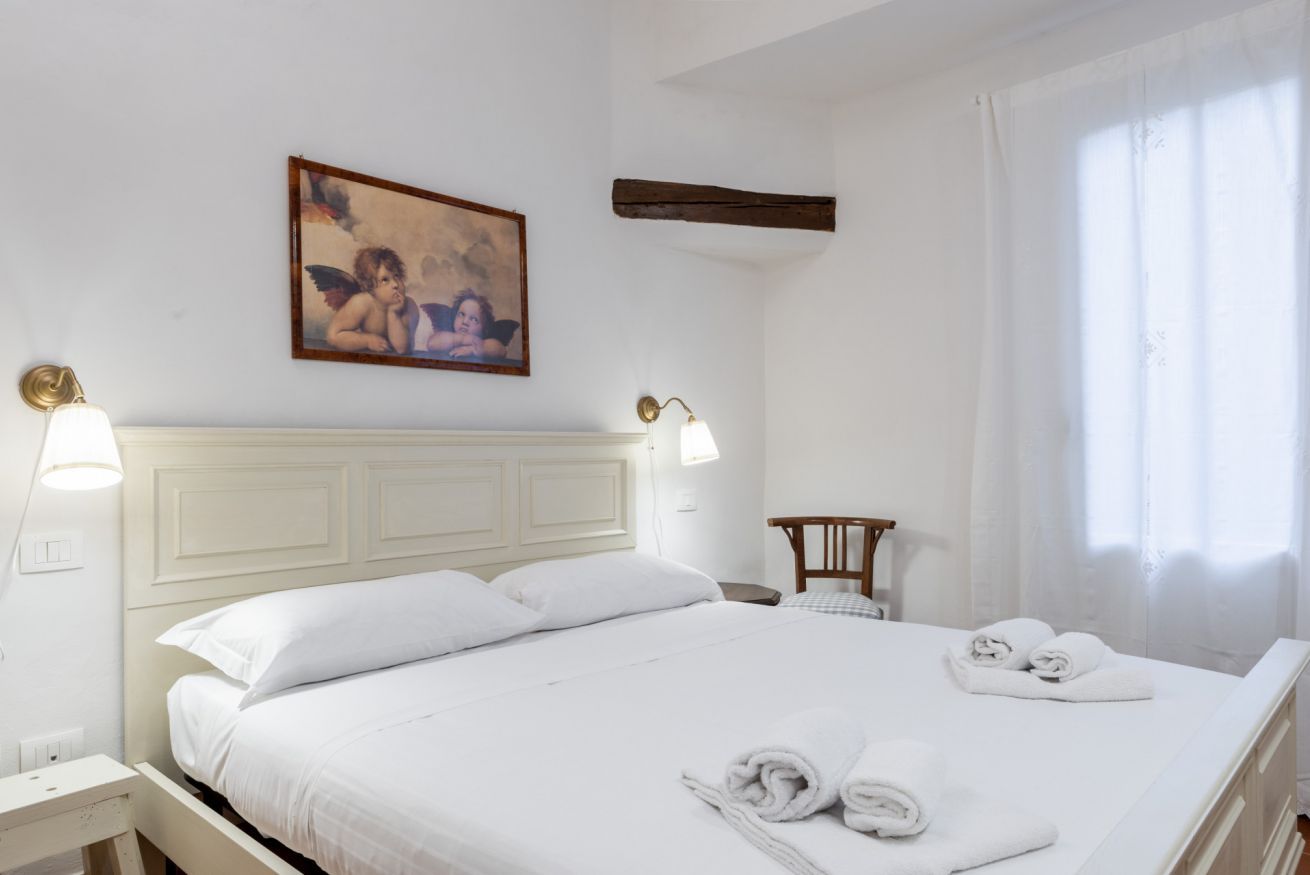 A neatly made bed with white linens and towels, a wooden headboard, a wall-mounted painting, and a window with curtains.