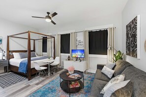 Una habitación de hotel moderna con una cama con dosel, un escritorio con silla, un sofá, una mesa de centro con revistas y un televisor.