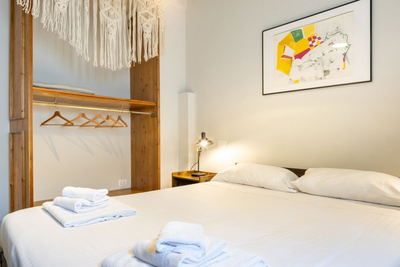 Une chambre à coucher comprenant un lit, une armoire en bois, une décoration murale en macramé, un tableau abstrait encadré et une table de chevet avec une lampe.