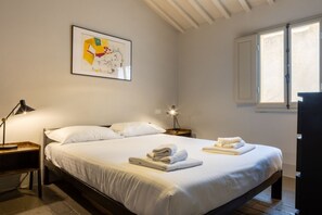 A neatly made bed with folded towels, a framed abstract painting on the wall, and a window with blinds. - CHIESA 12 3P - Florence (Firenze)