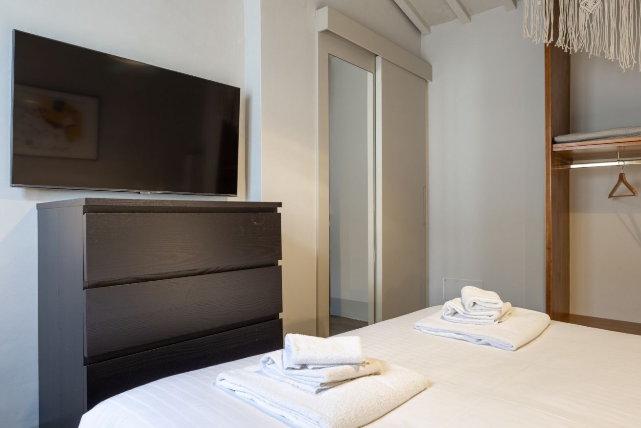 Una camera d'albergo moderna con un comò in legno scuro, una televisione a schermo piatto montata a parete, uno specchio grande e un letto rifatto con asciugamani piegati.