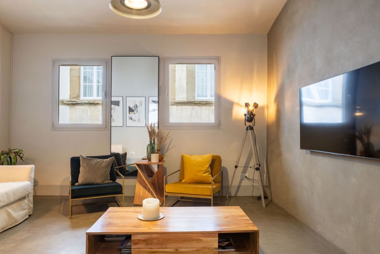 Un soggiorno moderno con un tavolino in legno, due poltrone, un divano, un grande specchio e una televisione a parete.