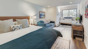 Ein modernes Schlafzimmer mit einem großen Bett, einem blauen Sofa, einem Schreibtisch mit Fernseher und einem Fenster mit Blick auf Bäume.
