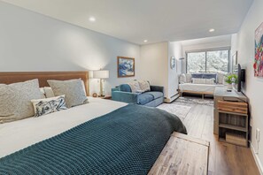 Ein modernes Schlafzimmer mit einem großen Bett, einem blauen Sofa, einem Schreibtisch mit Fernseher und einem Fenster mit Blick auf Bäume.