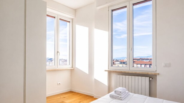 A bedroom with a bed, two windows, a radiator, and a view of buildings. - GuestHost - Cozy and modern 70 sqm apartment, able to comfortably accommodate up to 6 people, located on the 5th floor of a modern building with lift (NOT suitable for disabled people). The property is located in an extremely strategic area, just 400 met (Florence)