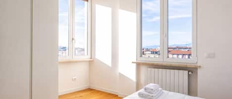 A bedroom with a bed, two windows, a radiator, and a view of buildings.