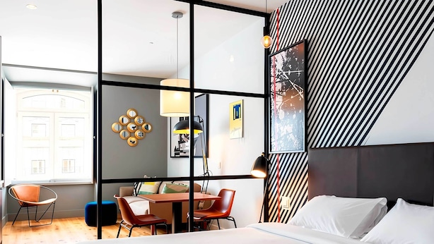 Moderni hotellihuone, jossa on suuri sänky, ruokailutila tuoleineen ja pöytineen sekä näkymä lasiovesta.