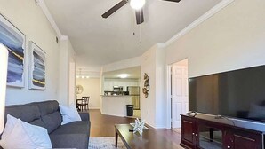 A living room with a sofa, coffee table, and TV stand. There is a ceiling fan, a kitchen in the background, and a dining area. - Walking Distance to Boutique Shops, Private Garage (Westchase)