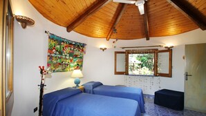 Ein Zimmer mit zwei Betten, Holzboden, Wandteppich in Farben, Lampe und Fenster mit Blick auf Grünflächen.