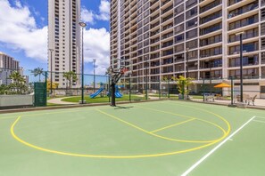Una cancha de baloncesto con aro, rodeada por una valla, y contigua a un edificio con varios balcones.