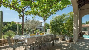An outdoor dining area with a table set for four, surrounded by chairs, under a pergola with climbing plants. - Casa Limonella - Maison 12 pers avec piscine (Fontvieille)