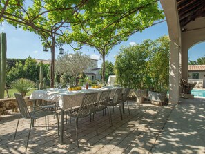 Un espacio de comedor al aire libre con una mesa preparada para cuatro personas, rodeada de sillas, bajo una pérgola con plantas trepadoras.
