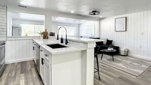 A modern kitchen with white cabinetry, a central island with a sink, and a dining area with a black chair and table. - Charming 3-bedroom vacation home near Siesta Key (Sarasota)