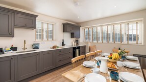 Una cocina moderna con armarios grises, una mesa de comedor de madera preparada para el desayuno y ventanas amplias con persianas.