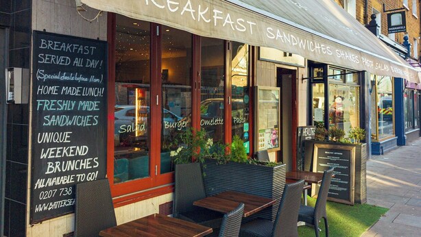 A breakfast cafe with outdoor seating, a signboard advertising breakfast sandwiches, and a nearby antique shop. - Homely flat by the river in Battersea, SW London - Pass the Keys (London)