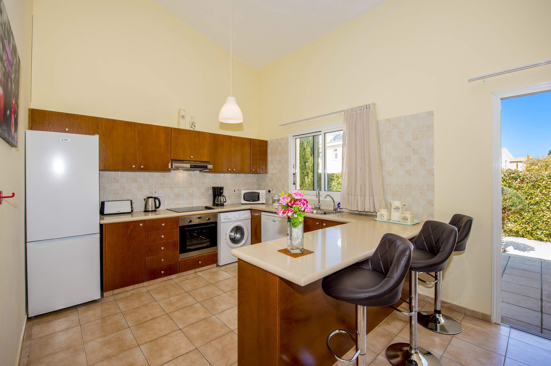 A modern kitchen with wooden cabinets, a built-in oven, a washing machine, and a dining area with two bar stools.