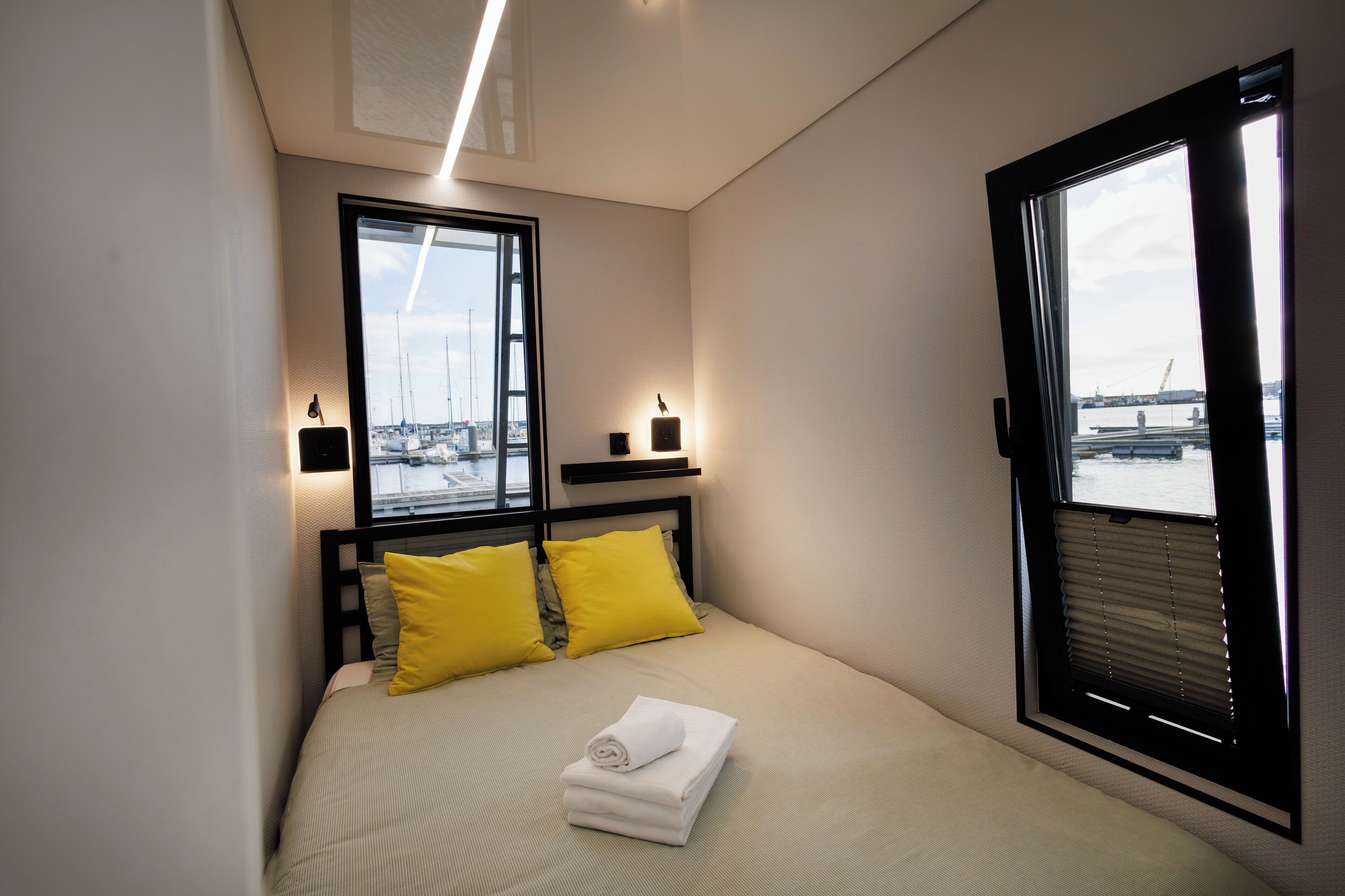 A neatly made bed with two pillows, a folded towel, and a view of a marina through a window.