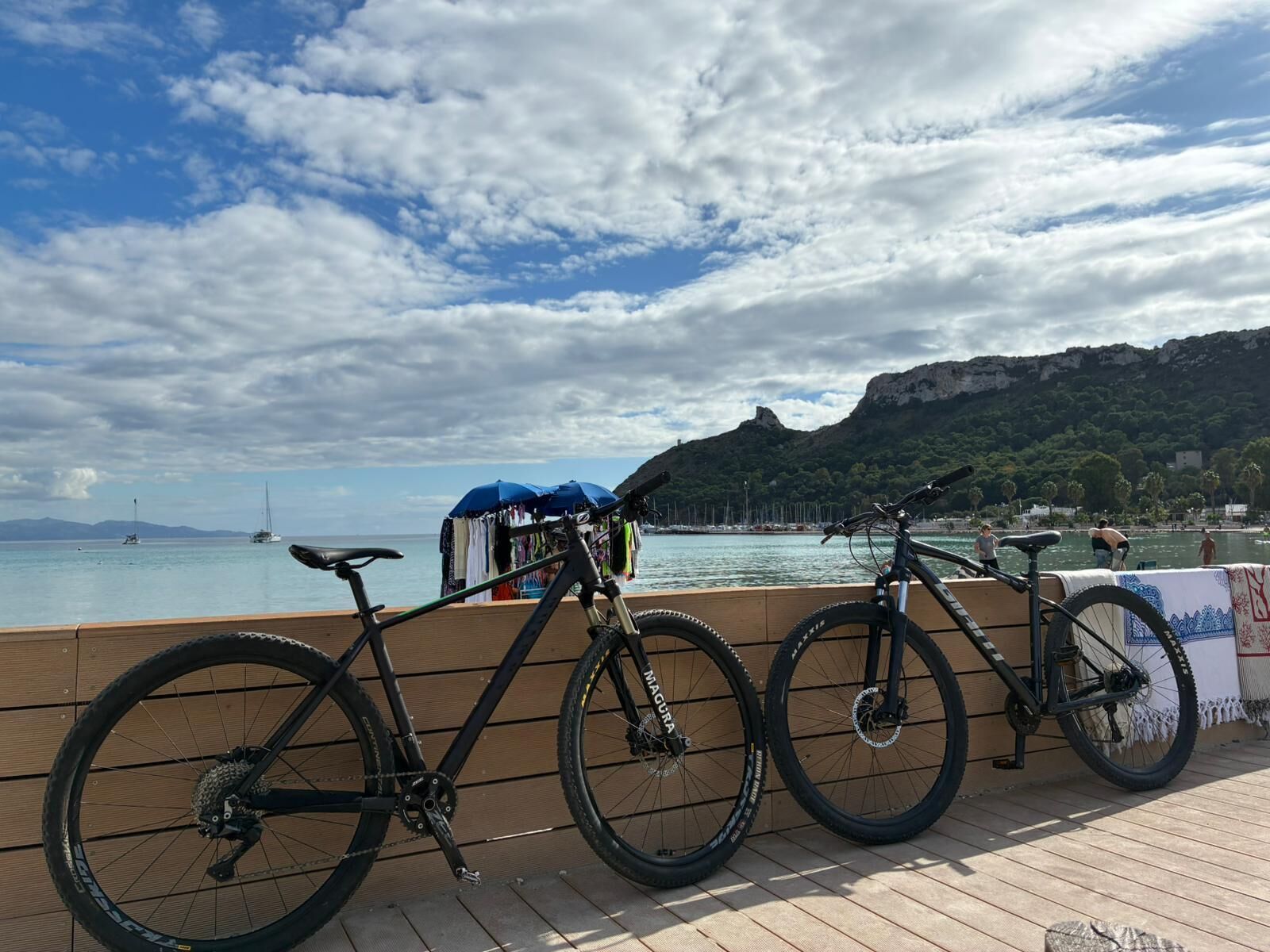 A wooden deck with bicycles parked, overlooking a body of water with boats and a mountainous backdrop.