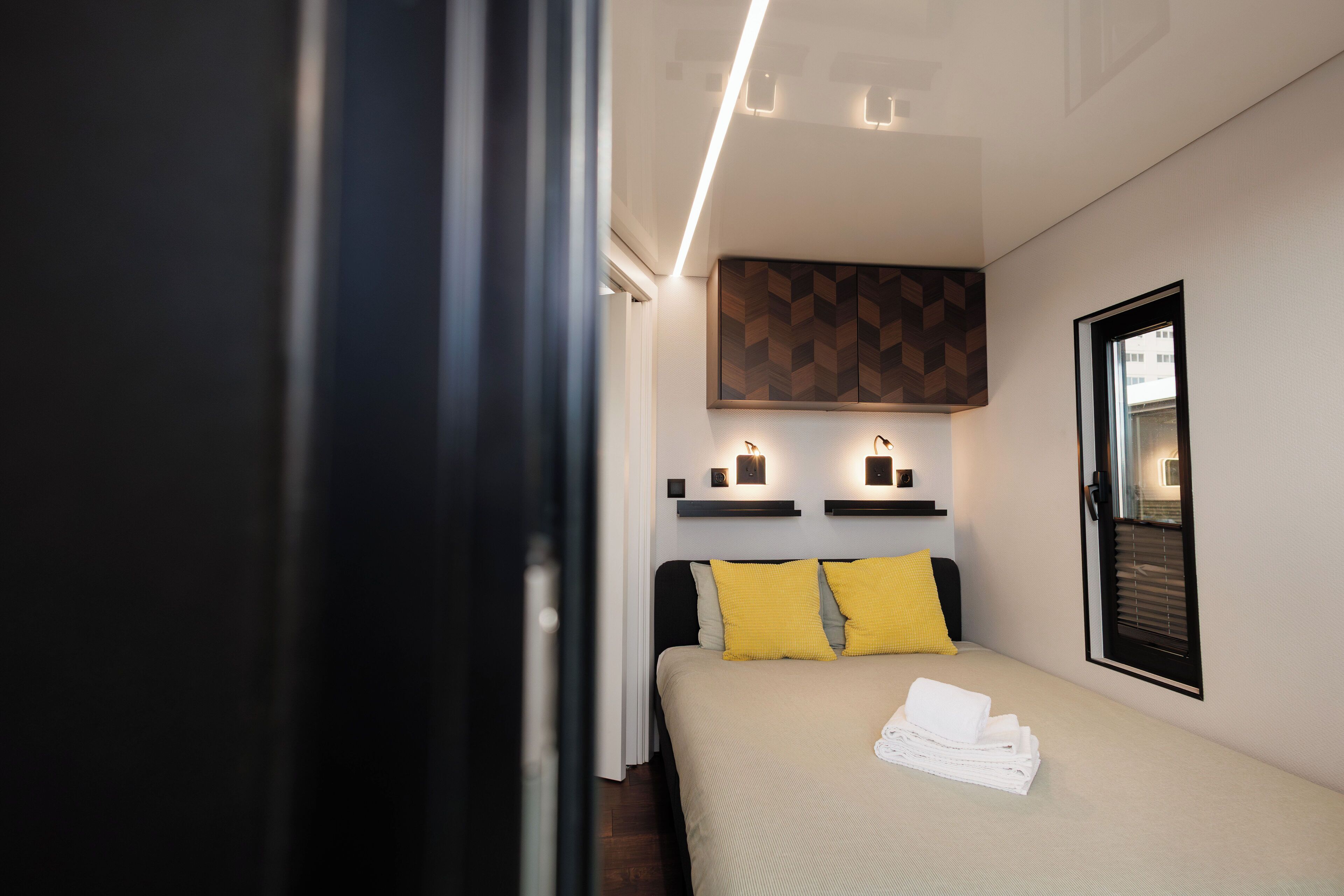 Et moderne hotelværelse med en pænt redt seng, en vægmonteret hylde med stearinlys og et vindue med rullegardiner.