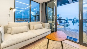A modern living room with a sofa, a small wooden table, and large windows offering a view of a marina. - The Homeboat Company Sant'Elmo-Cagliari (Cagliari)