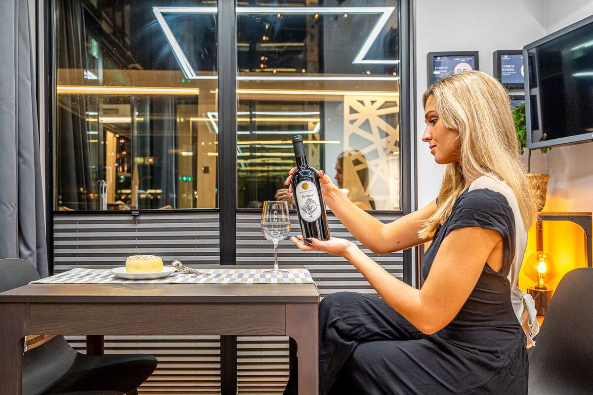 A woman holding a wine bottle at a table with a glass, a plate with a dessert, and a lamp in a modern interior with a geometric wall design.
